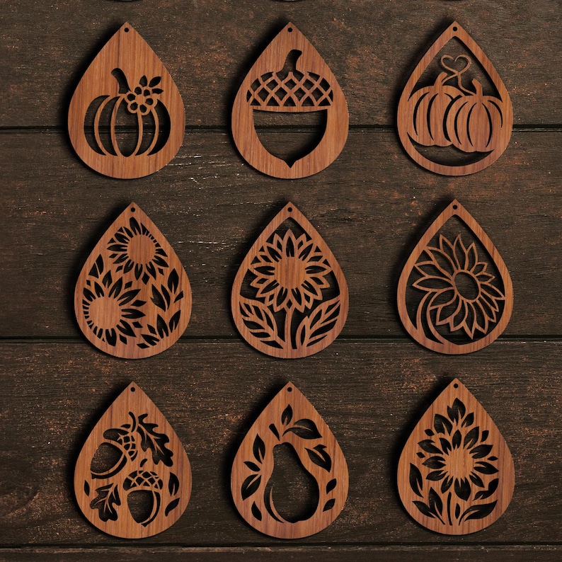 K&ouml;nnte beinhalten: Neun holzfarbene Ohrringe in Tropfenform mit ausgeschnittenen Designs. Die Designs umfassen K&uuml;rbisse, Eicheln, Sonnenblumen und eine Birne. Die Ohrringe haben eine warme braune Farbe und sind auf einer dunklen Holzoberfl&auml;che angeordnet.