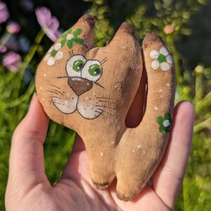 Könnte beinhalten: Ein braunes Stoffkatzenspielzeug mit weißen und grünen Blumenakzenten. Die Katze hat ein bemaltes Gesicht mit großen Augen und einer braunen Nase.