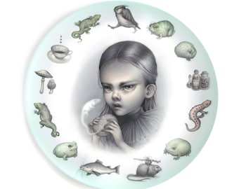May include: A round ceramic plate featuring a central illustration of a stern-faced child holding a small white creature. The border is decorated with various illustrations including a teacup, mushrooms, lizards, frogs, a fish, and glass bottles.