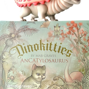 May include: A collectible figurine of a Dinokittie, an Ancatylosaurus, with a pink and white shell-like back and a cat-like face. The figurine sits atop a box with illustrations of Dinokitties and the text "Dinokitties by Mab Graves Ancatylosaurus".