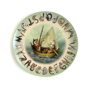 May include: A round decorative plate with a light green border featuring the alphabet letters A through Z, each accompanied by a small cat illustration. The center depicts an owl playing a violin and a cat rowing a sailboat on the water.