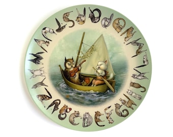 May include: A round decorative plate with a light green border featuring the alphabet letters A through Z, each accompanied by a small cat illustration. The center depicts an owl playing a violin and a cat rowing a sailboat on the water.