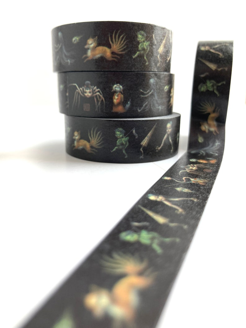 May include: A roll of black washi tape with a repeating pattern of colourful illustrations of fantastical creatures, including a fox, a spider, and a goblin. The tape is wound around three spools.
