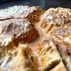 May include: Close-up of a golden-brown, crusty baked good, possibly bread or pastry, cut into wedges. The surface has a textured, uneven appearance with visible layers and crevices. The lighting highlights the details of the baked item.