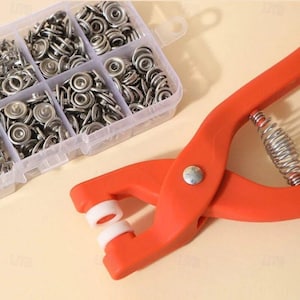 May include: A clear plastic organizer filled with silver metal snap fasteners. An orange snap fastener tool with white plastic tips and a silver spring is in the foreground. The tools are for sewing and crafting.