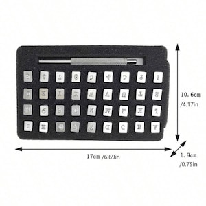 Może przedstawiać: Czarne etui z zestawem 40 metalowych stempli z literami i cyframi. Etui ma 17 cm szerokości i 10,6 cm wysokości. Stempelki są ułożone w rzędach i kolumnach. W zestawie znajduje się metalowe narzędzie do stemplowania.