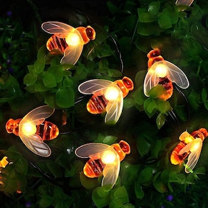 May include: String of solar-powered fairy lights shaped like bees with warm white LED lights. The lights are strung on a black wire and are perfect for adding a touch of whimsy to any outdoor space.