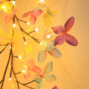K&ouml;nnte beinhalten: Lichterkette mit bunten Schmetterlings-Dekorationen. Die Lichter sind warmwei&szlig; und die Schmetterlinge sind in den Farben Rosa, Lila, Blau und Gelb.