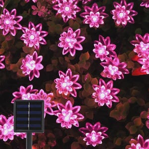 May include: Pink solar-powered flower lights. The lights are shaped like flowers with multiple petals and a bright center. The lights are arranged among green foliage, with a solar panel in the foreground. The lights are a vibrant pink color.