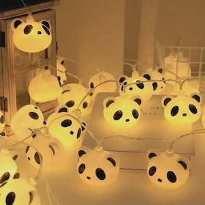 Könnte beinhalten: Lichterkette mit Panda-Formen. Jede Leuchte ist weiß mit schwarzen Augen und Ohren. Die Lichter sind an einem dünnen, transparenten Draht aufgereiht. Die Lichter leuchten und erzeugen einen warmen Schein.