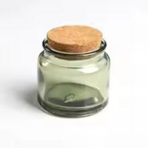 May include: A small, clear glass jar with a round cork lid. The jar has a subtle green tint and a wide opening. The cork lid is circular and fits snugly. The jar is on a white surface.
