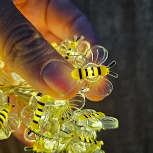 May include: A string of fairy lights with clear plastic bee-shaped bulbs. The bees have yellow bodies with black stripes and clear wings.