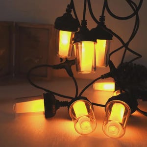 May include: String of outdoor string lights with clear glass shades and warm yellow LED bulbs. The black cords and fixtures create a modern look. The lights are arranged on a light-colored surface.