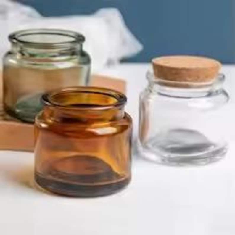 May include: Three glass jars of different sizes and colours. One is amber, one is clear, and the third is a light green. The clear jar has a cork stopper. The jars are on a white surface with a blue background.