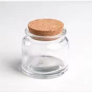 May include: A small, clear glass jar with a wide opening and a round, natural cork stopper. The jar is empty and sits on a white background. The cork is a light brown colour, contrasting with the transparent glass.