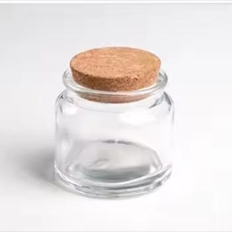 May include: A small, clear glass jar with a wide opening and a round, natural cork stopper. The jar is empty and sits on a white background. The cork is a light brown colour, contrasting with the transparent glass.