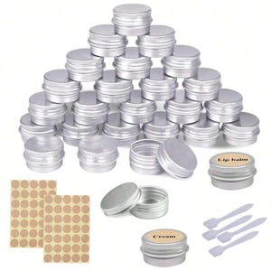 Op de afbeelding: Een set van 24 zilveren aluminium mini-blikken met deksels. De blikken zijn in een piramidevorm gestapeld. Er zijn ook twee open blikken, een met het label "Lip balm" en de andere met het label "Cream". De set bevat ook drie kleine plastic spatels en een vel met 100 ronde stickers.