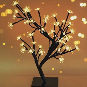 Puede incluir: Un árbol decorativo negro con luces en forma de flor. El árbol tiene una base cuadrada y un tronco curvo con múltiples ramas. Las luces son de color amarillo cálido, creando un brillo suave. El fondo es un degradado de naranja y amarillo.