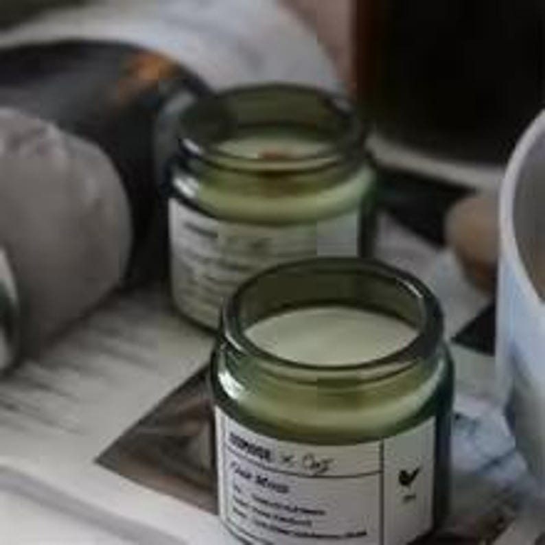 May include: Two small, green-tinted glass jars filled with white wax candles. Each jar has a white label with black text. The jars sit on a printed page, with a blurred background including a white mug and other objects.