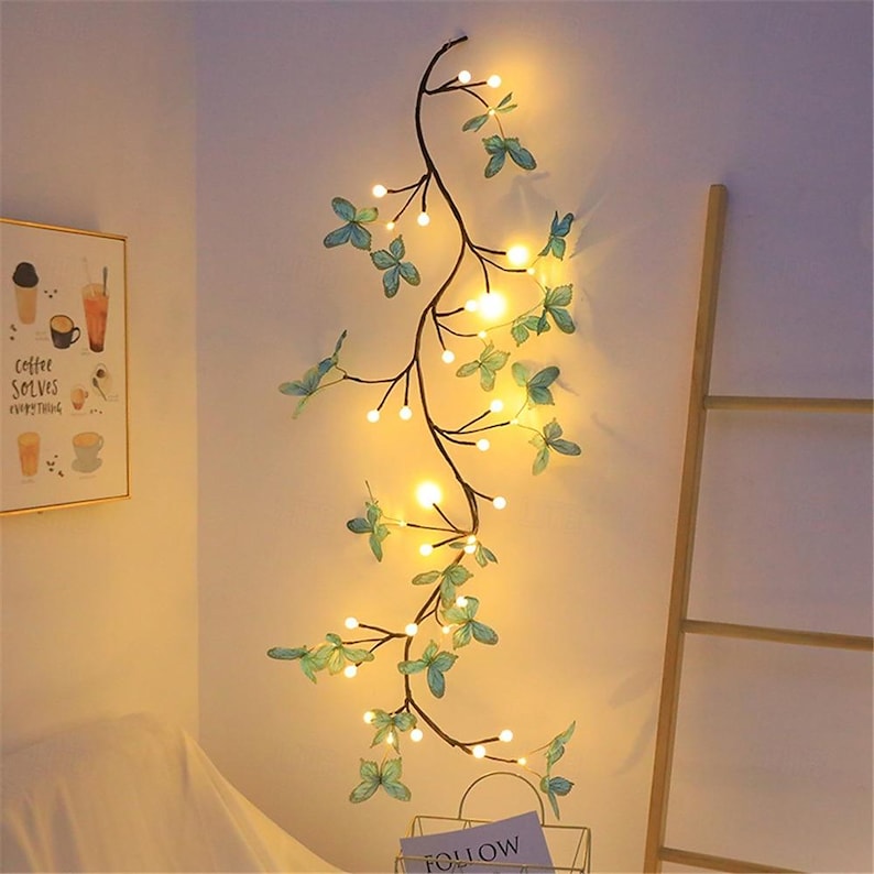 K&ouml;nnte beinhalten: Eine Lichterkette mit kleinen wei&szlig;en Gl&uuml;hbirnen und gr&uuml;nen Bl&auml;ttern in Form von Schmetterlingen. Die Lichter sind auf einem schwarzen Draht aufgef&auml;delt und h&auml;ngen an einer wei&szlig;en Wand.