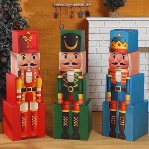 Op de afbeelding: Drie kleurrijke, gestapelde notenkrakerfiguren. De notenkrakers zijn rood, groen en blauw, met gedetailleerde illustraties van uniformen, snorren en hoeden. De figuren staan voor een open haard en een kerstboom.