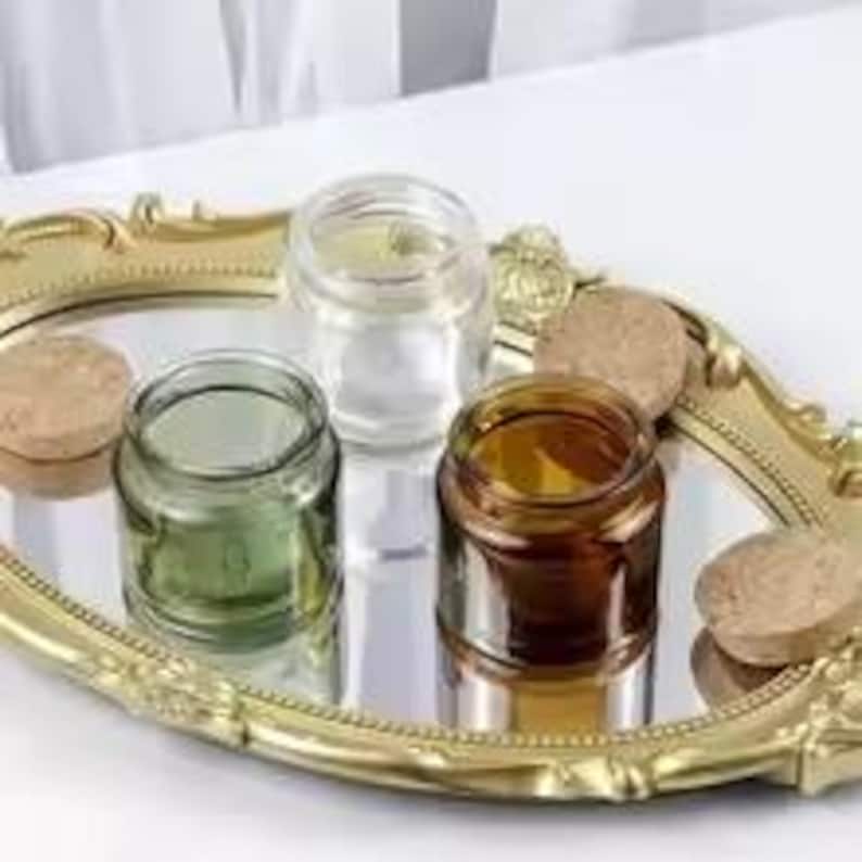 May include: Three small glass jars, one green, one clear, and one amber, sit on a gold-coloured, ornate tray. Cork lids are placed beside the jars. The jars are reflected in the mirrored surface of the tray.