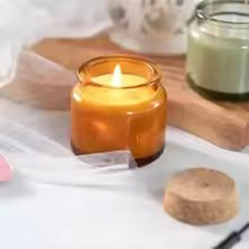 May include: An amber-coloured candle burning in a small glass jar. The candle is on a white surface with a wooden board and a small wooden lid. Another candle in a jar is visible in the background.