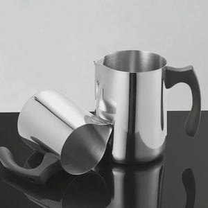 May include: Two stainless steel milk frothing pitchers with black handles. One pitcher is upright, and the other is on its side. The pitchers have a shiny, reflective surface and a spout for pouring.