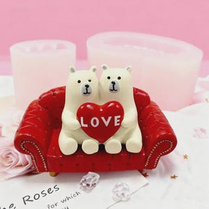 May include: A figurine of two white polar bears sitting on a red couch, holding a red heart with the word "LOVE" on it. The bears have black eyes and noses. The couch has a tufted design.