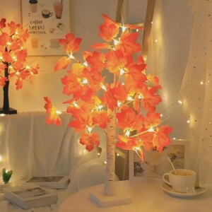 Könnte beinhalten: Ein dekorativer Baum mit orangefarbenen Ahornblättern und kleinen Lichtern. Der Baum hat einen weißen Stamm und steht auf einem quadratischen Sockel. Die Blätter sind beleuchtet und erzeugen einen warmen Schein. Herbstliche Dekoration.