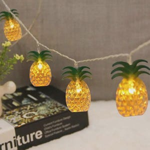 May include: A string of decorative lights featuring pineapple-shaped bulbs. Each pineapple has a textured, yellow-orange body and green leafy top. The lights are strung on a white wire, creating a tropical and festive ambiance.