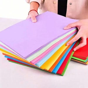 Op de afbeelding: Een stapel kleurrijke papieren vellen in diverse tinten, waaronder paars, roze, blauw, geel, oranje en rood. Het papier wordt door handen vastgehouden, wat de levendige kleuren laat zien. Ideaal voor knutselen, kunstprojecten of kantoorgebruik.