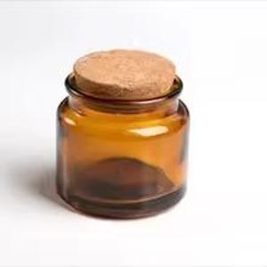 May include: A small, amber glass jar with a cork stopper. The jar has a rounded shape and a wide opening. The cork is circular and fits snugly on top. The jar is empty and sits on a white background.