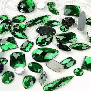 May include: An assortment of faceted green gemstones in various shapes and sizes, including teardrops, ovals, and squares. Some have a metallic backing, while others are clear. The gems are scattered on a white, textured surface.