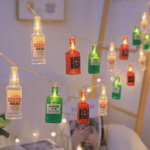 Puede incluir: Una cadena de luces LED con botellas de plástico en miniatura de varios colores, incluyendo transparente, verde, rojo y naranja. Cada botella contiene una pequeña luz y presenta una etiqueta con texto como VODKA YOUR LABEL HERE y LONDON GIN.