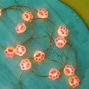 May include: String of decorative lights featuring paw prints. Each paw print is a soft pink color with a darker pink paw pad design. The lights are strung on a thin, gold-colored wire. The background is a teal-colored, textured surface.