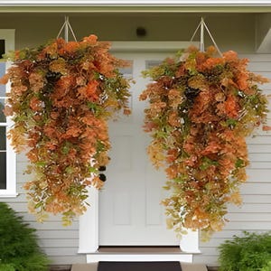 Peut inclure: Deux paniers suspendus remplis de feuillage d'automne artificiel. Les compositions présentent un mélange de feuilles et de fleurs orange, marron et vertes, créant une ambiance chaleureuse et automnale. Les paniers sont suspendus à un porche blanc, ajoutant une touche décorative saisonnière.