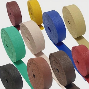 Könnte beinhalten: Eine Sammlung von zehn Rollen strukturiertem Gurtband in verschiedenen Unifarben, darunter Schwarz, Grün, Kastanienbraun, Gelb, Beige, Blau, Hellbraun, Dunkelbraun und Rot. Jede Rolle ist fest gewickelt und weist eine gleichmäßige Breite sowie eine körnige Oberfläche auf.