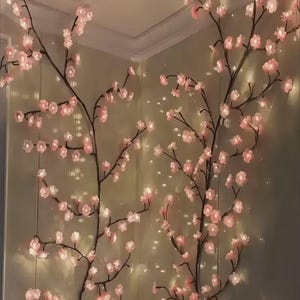 以下が含まれることがあります： 柔らかなピンク色の花と温白色のライトが付いた、人工の桜の枝。枝は濃い茶色で、繊細で照らされたディスプレイを作り出しています。花は枝に沿って集まっており、ロマンチックでエレガントな雰囲気を醸し出しています。