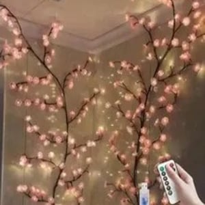 Puede incluir: Ramas decorativas iluminadas de flores de cerezo con flores rosas y luces blancas cálidas. Las ramas están dispuestas en una esquina, creando un brillo suave. Se sostiene un mando a distancia en la mano.