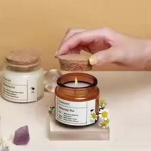 May include: A lit candle in a small, round, amber glass jar with a cork lid. The jar sits on a clear acrylic block, with a label and small white flowers. Another jar with a cork lid is in the background.