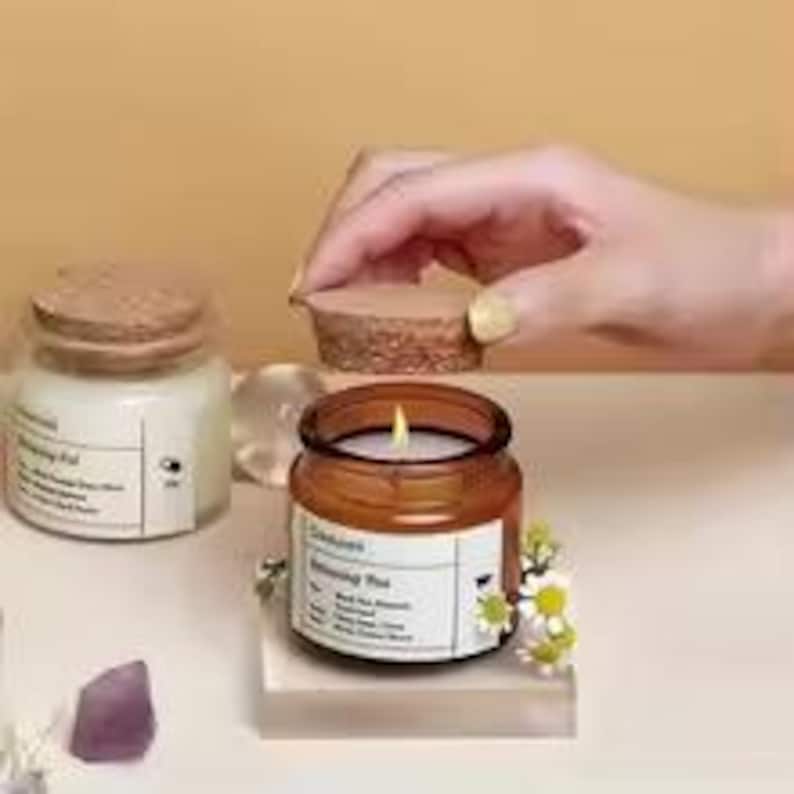 May include: A lit candle in a small, round, amber glass jar with a cork lid. The jar sits on a clear acrylic block, with a label and small white flowers. Another jar with a cork lid is in the background.