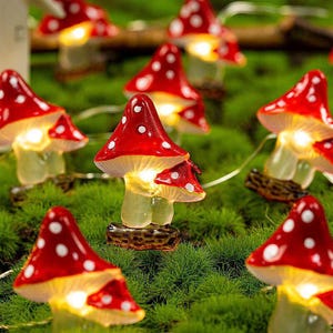 May include: A string of decorative lights featuring red mushroom-shaped lamps with white polka dots. The mushroom lights are illuminated with a warm glow, set against a backdrop of green moss. The lights are connected by a thin wire.