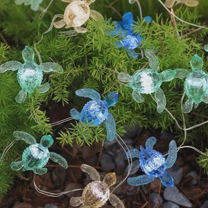 Puede incluir: Una cadena de luces decorativas con formas de tortugas marinas translúcidas en tonos azules, verdes y ámbar. Las tortugas están ensartadas en un alambre fino y transparente y están dispuestas entre follaje verde y pequeñas rocas.