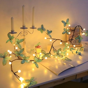 May include: A string of fairy lights with butterfly accents and warm white bulbs. The lights are wrapped around a branch and are illuminated against a white background.