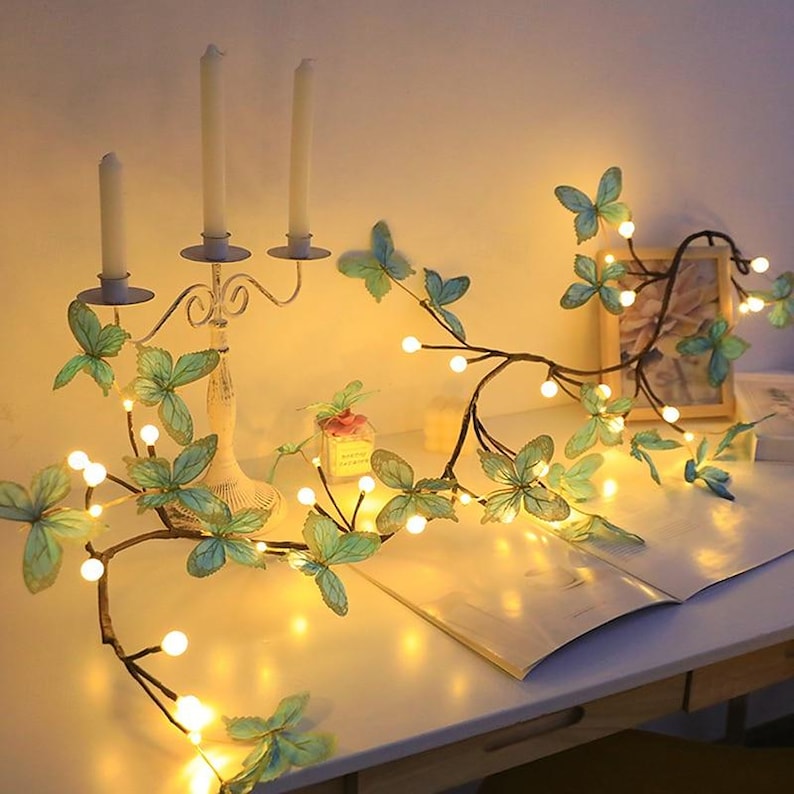 K&ouml;nnte beinhalten: Eine Lichterkette mit Schmetterlingsakzenten und warmwei&szlig;en Gl&uuml;hbirnen. Die Lichter sind um einen Ast gewickelt und leuchten vor einem wei&szlig;en Hintergrund.