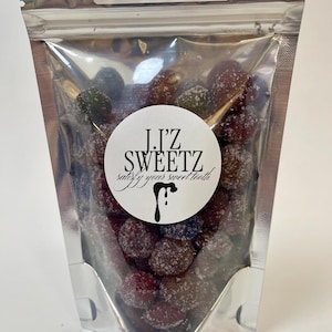 Puede incluir: Una bolsa plateada resellable llena de caramelos azucarados rojos y azules. La bolsa tiene una etiqueta blanca que dice "Gracias". Una pegatina circular blanca con texto negro dice "J.I'Z SWEETZ satisfy your sweet tooth" con un diseño de caramelo negro que gotea.