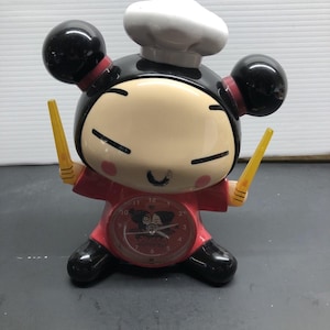 May include: A novelty alarm clock featuring a cartoon character in a chef's hat and red outfit. The clock has a round face with a cartoon graphic and yellow chopsticks. The character's hair is black with red accents.