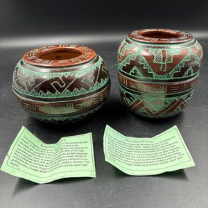 May include: Two round, dark brown ceramic pots with teal geometric patterns etched into the surface. Each pot has a wide opening and a short, rounded body. Two light green paper tags with black text describing Brazilian pottery are included.