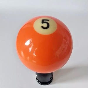May include: Orange billiard ball with a white number 5 on it. The ball is attached to a black base.